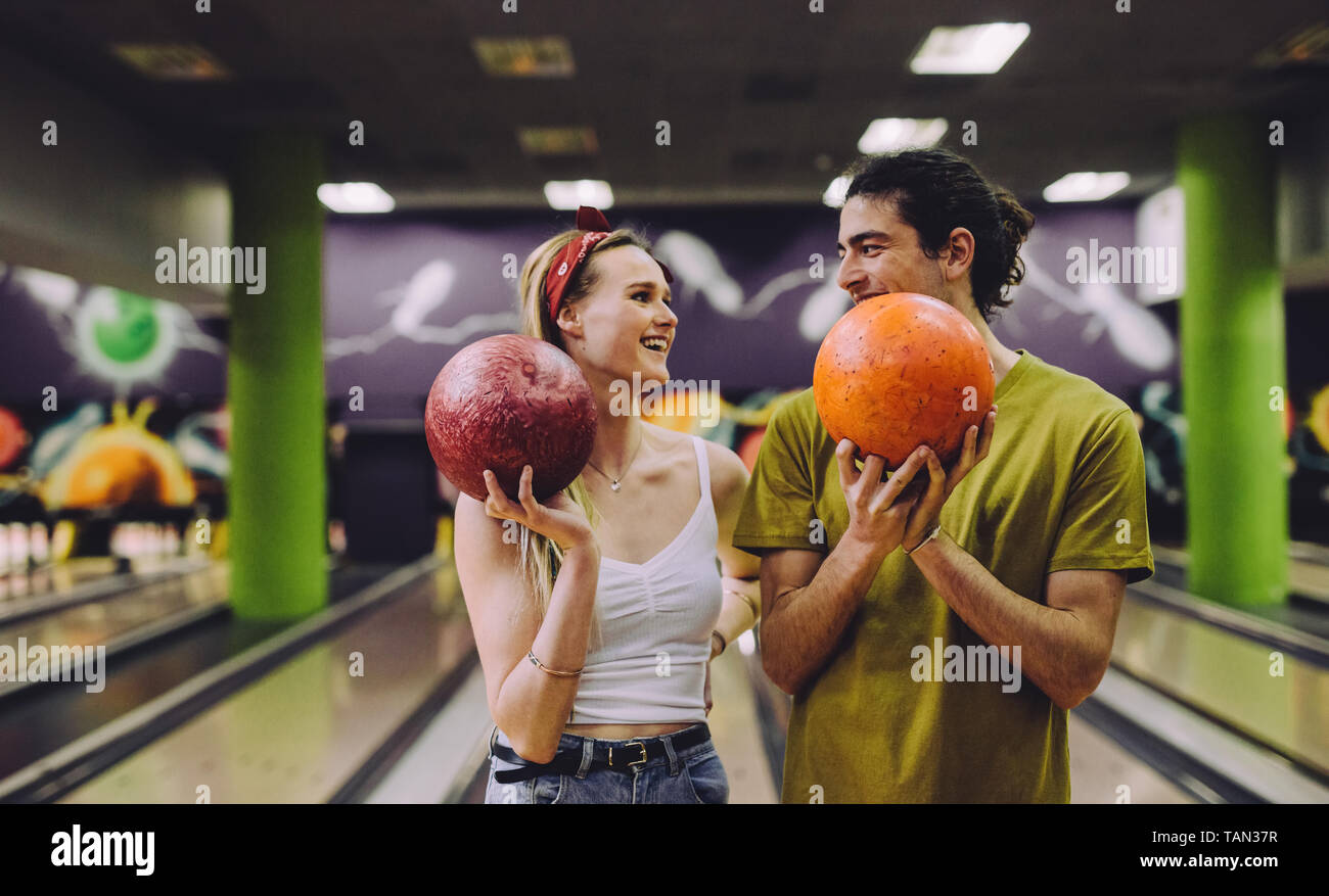 Fröhlicher junger Paare auf der Bowlingbahn mit den Kugeln an jedem anderen suchen. Junger Mann und Frau, die in einem bowling club Holding Kugeln. Stockfoto