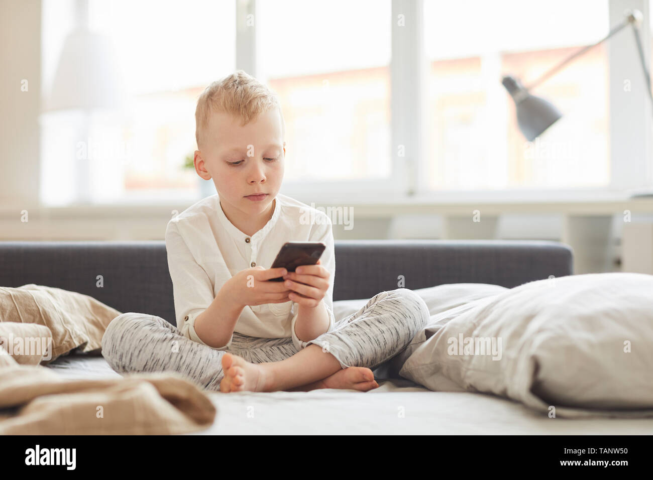 Ernsthafte konzentriert blonde Junge im homewear Sitzen mit gekreuzten Beinen auf dem Bett und Sie ihr Smartphone im Morgen Stockfoto