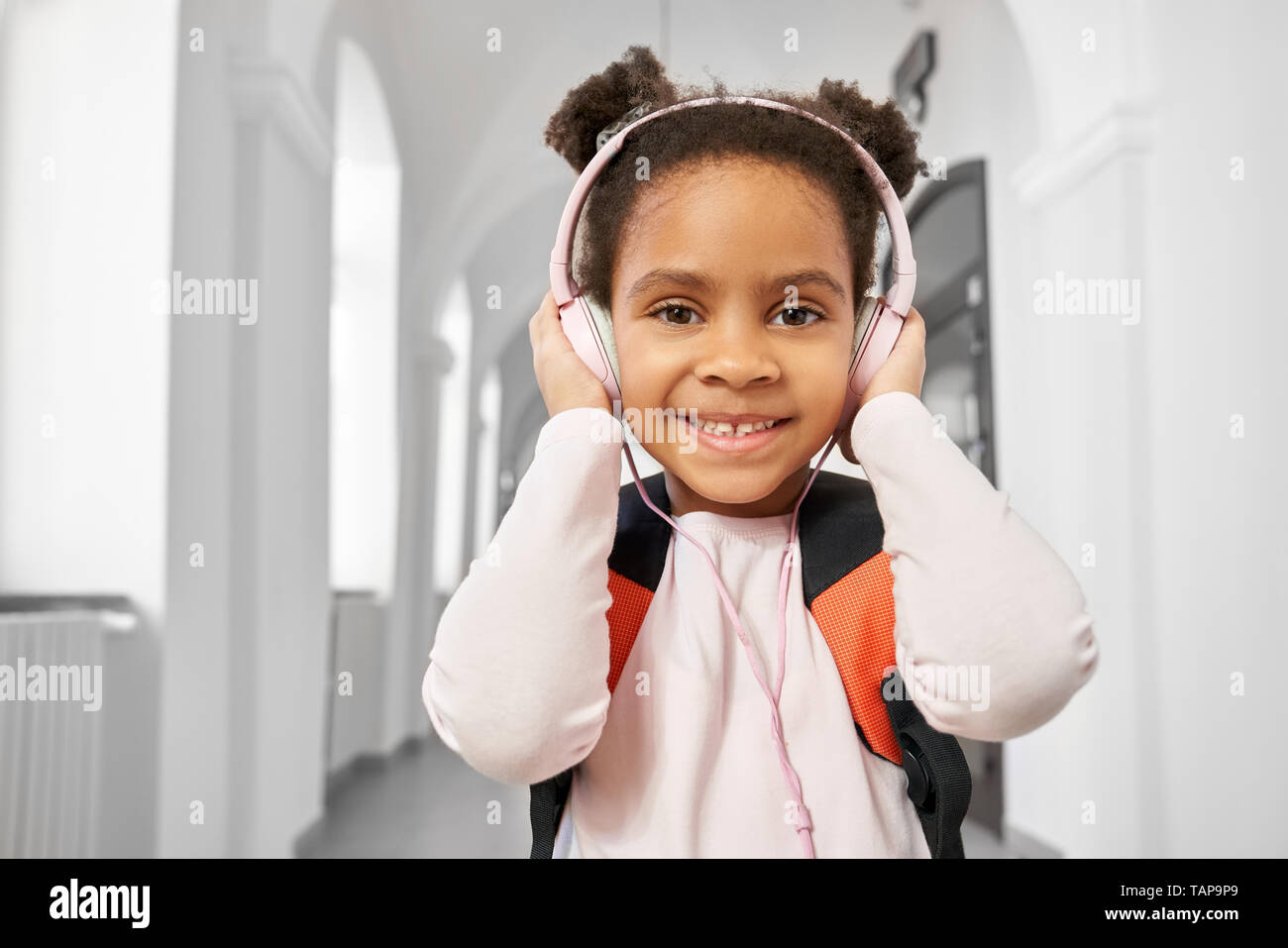 Positive, schöne, freundliche Schulmädchen mit Kopfhörern auf dem Flur der Schule prrimary posieren. Hübsches, lächelnde Mädchen hören Musik in der Pause zwischen den Unterrichtsstunden. Stockfoto