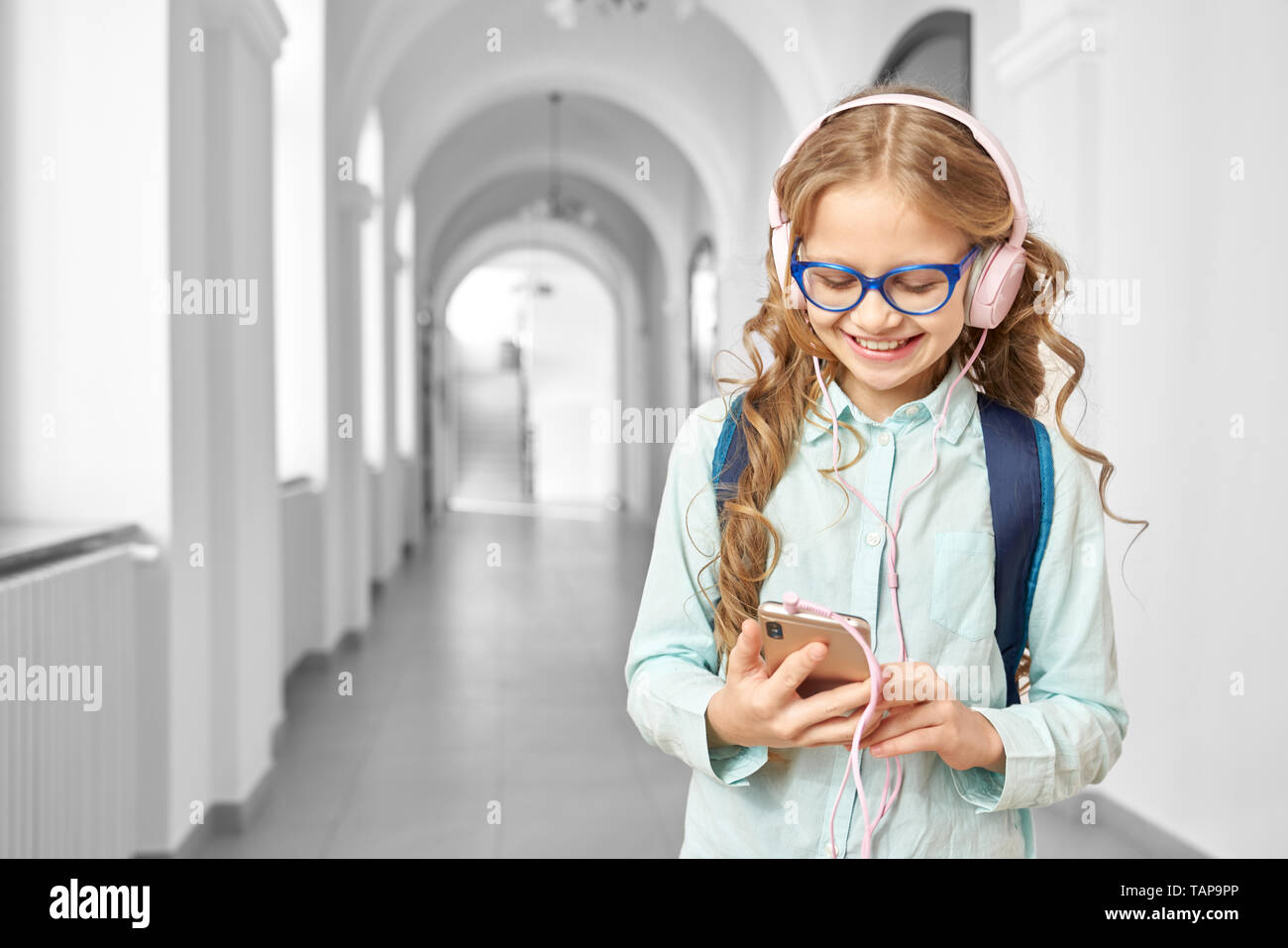 Schön, schöne Schule Mädchen Musik hören mit Kopfhörern in der Pause zwischen den Unterrichtsstunden. Happy girl in Gläsern, Telefon, lächelnd und stand auf dem Flur der Schule. Stockfoto