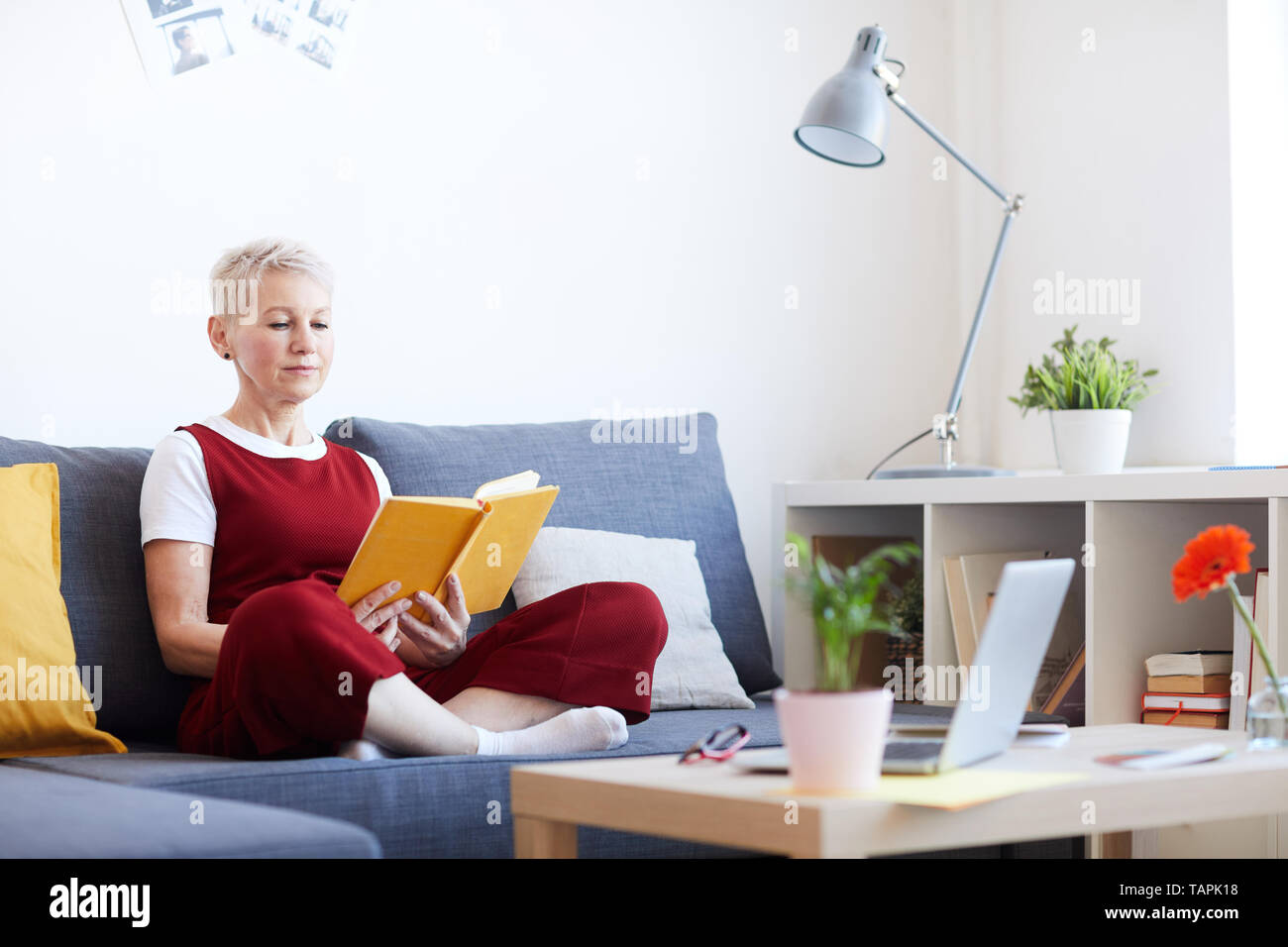 Blonde Frau mittleren Alters in der casualwear sitzen auf der Couch und Lesen interessante Buch der Geschichten in der Freizeit Stockfoto