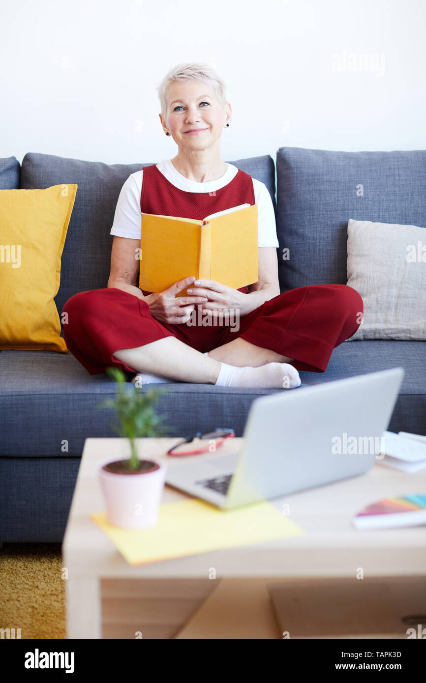 Gerne blonde Frau in der casualwear entspannen auf der Couch mit Buch beim Verbringen von Wochenende zu Hause Stockfoto