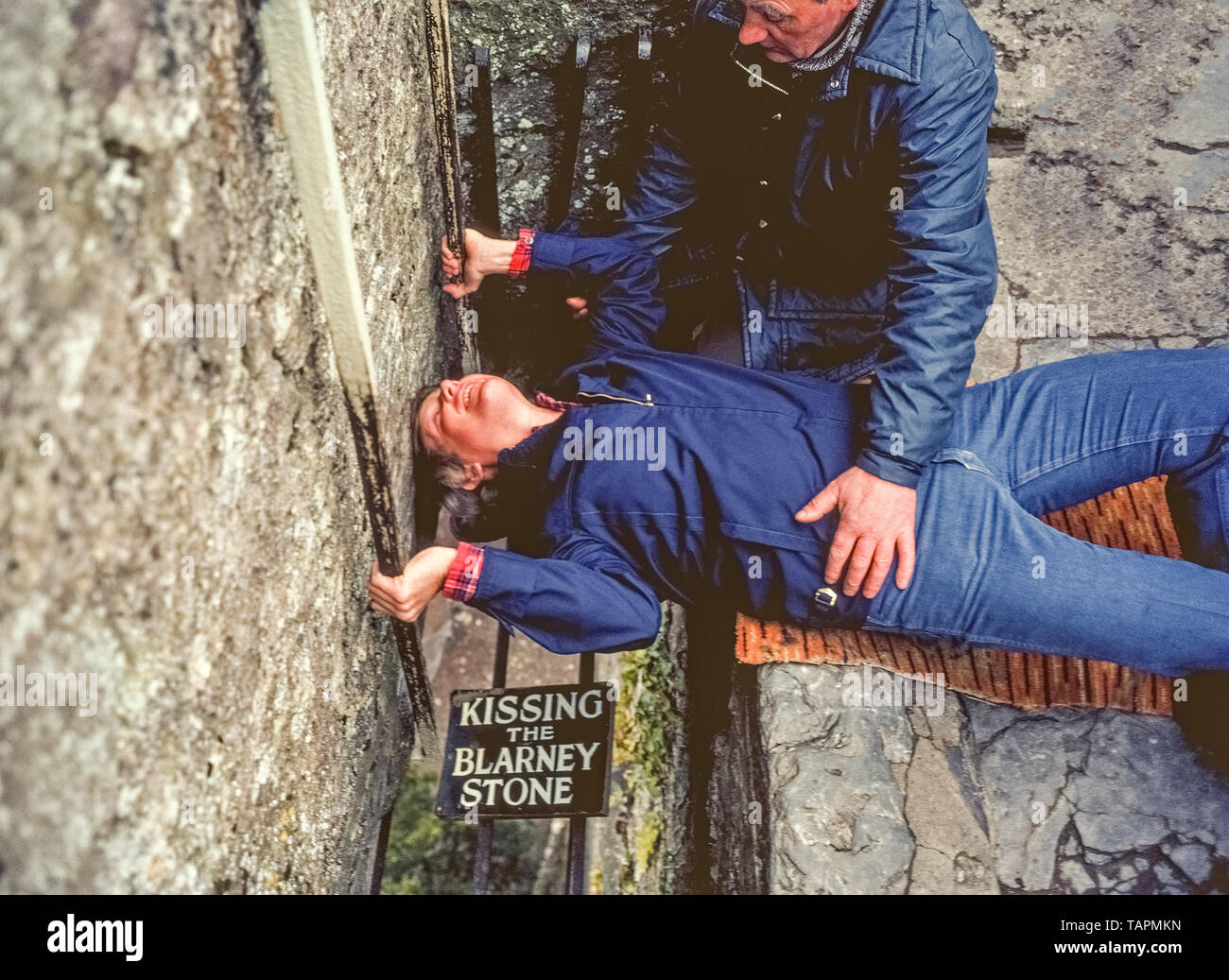 Eine Telefonzentrale hält auf der jungen amerikanischen Mann, der auf dem Rücken liegt, packt zwei Metall Geländer, und lehnt seinen Kopf zurück in den traditionellen upside-down Art und Weise den berühmten Blarney Stone hoch in den Turm von Blarney Castle im County Cork, Irland zu küssen. Es hat eine lange Tradition für die Besucher des Schloss aus dem 15. Jahrhundert wurden die schmalen Treppe der berühmte Satz von Kalkstein auf den Zinnen, die vom Turm aus zu erreichen, um zu klettern. Bügeleisen bars verhindern die Münder von Fallen durch den offenen Raum auf den Boden. Küssen der Stein sagte Beredsamkeit zu verleihen; in anderen Worten, geben ihnen das Geschenk der Akv. Stockfoto