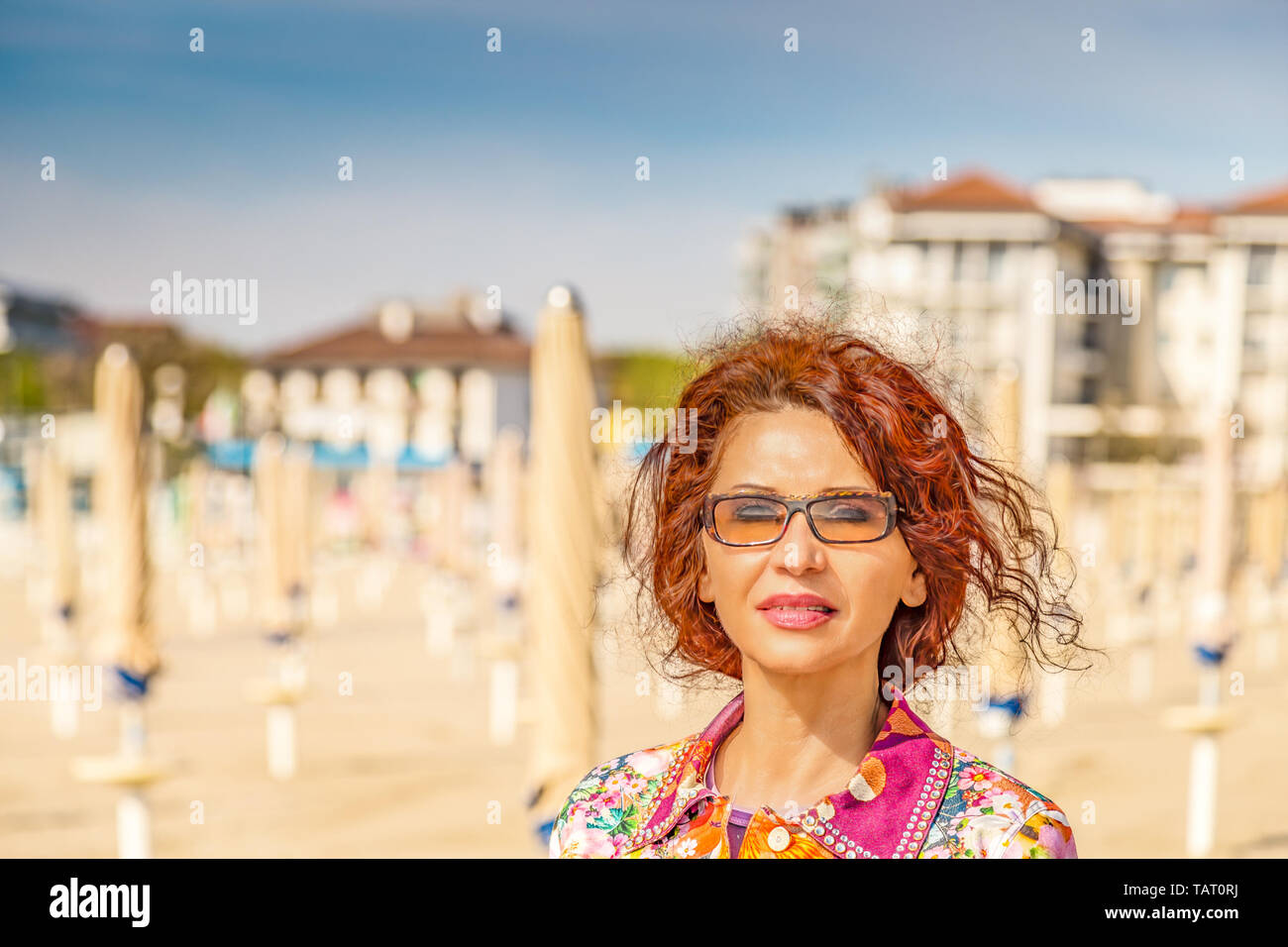 Nahaufnahme der mediterran bepflanzten Frau mit Gipsy rotes Haar und gebräunte Haut tragen bunte luxuriöses Hemd und braune Sonnenbrille lächelnd am Meer Stockfoto