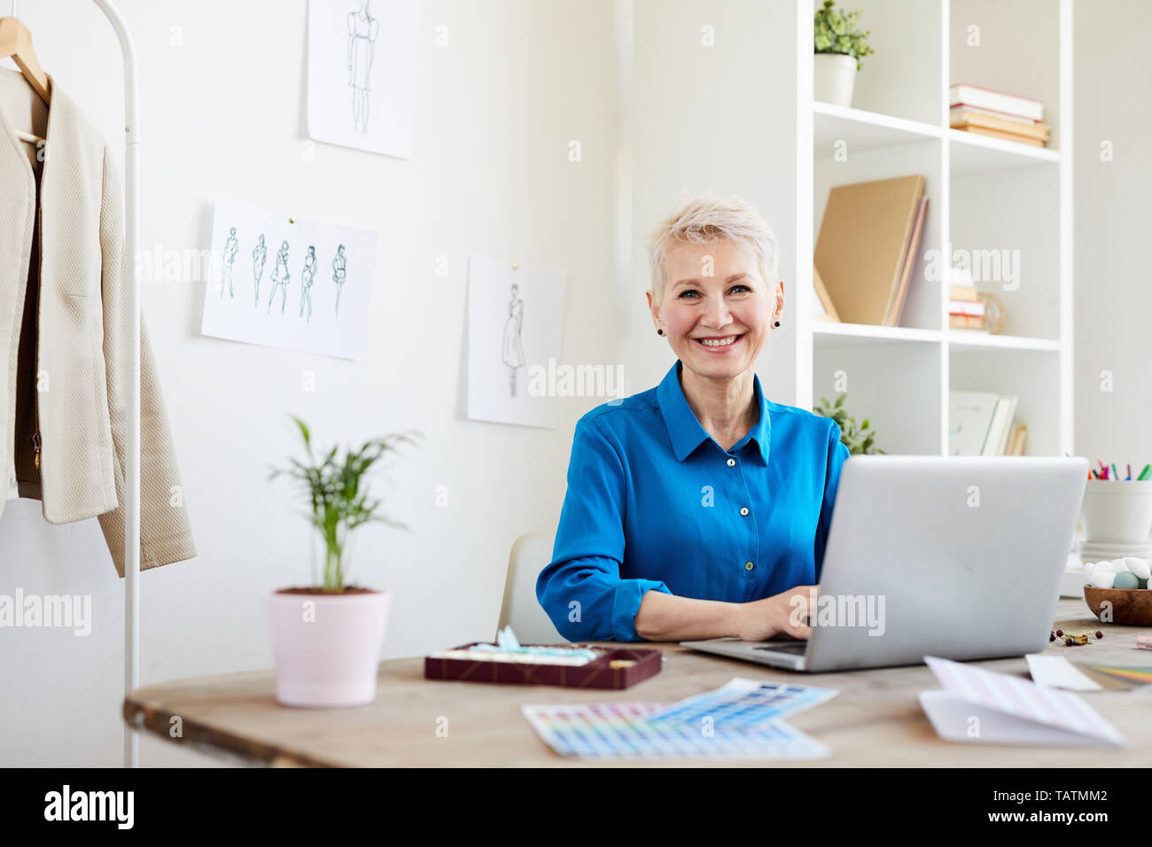 Fröhliche reife blonde Frau in Smart Casual sitzend durch Tisch vor Laptop beim Surfen über Websites für neue Ideen Stockfoto