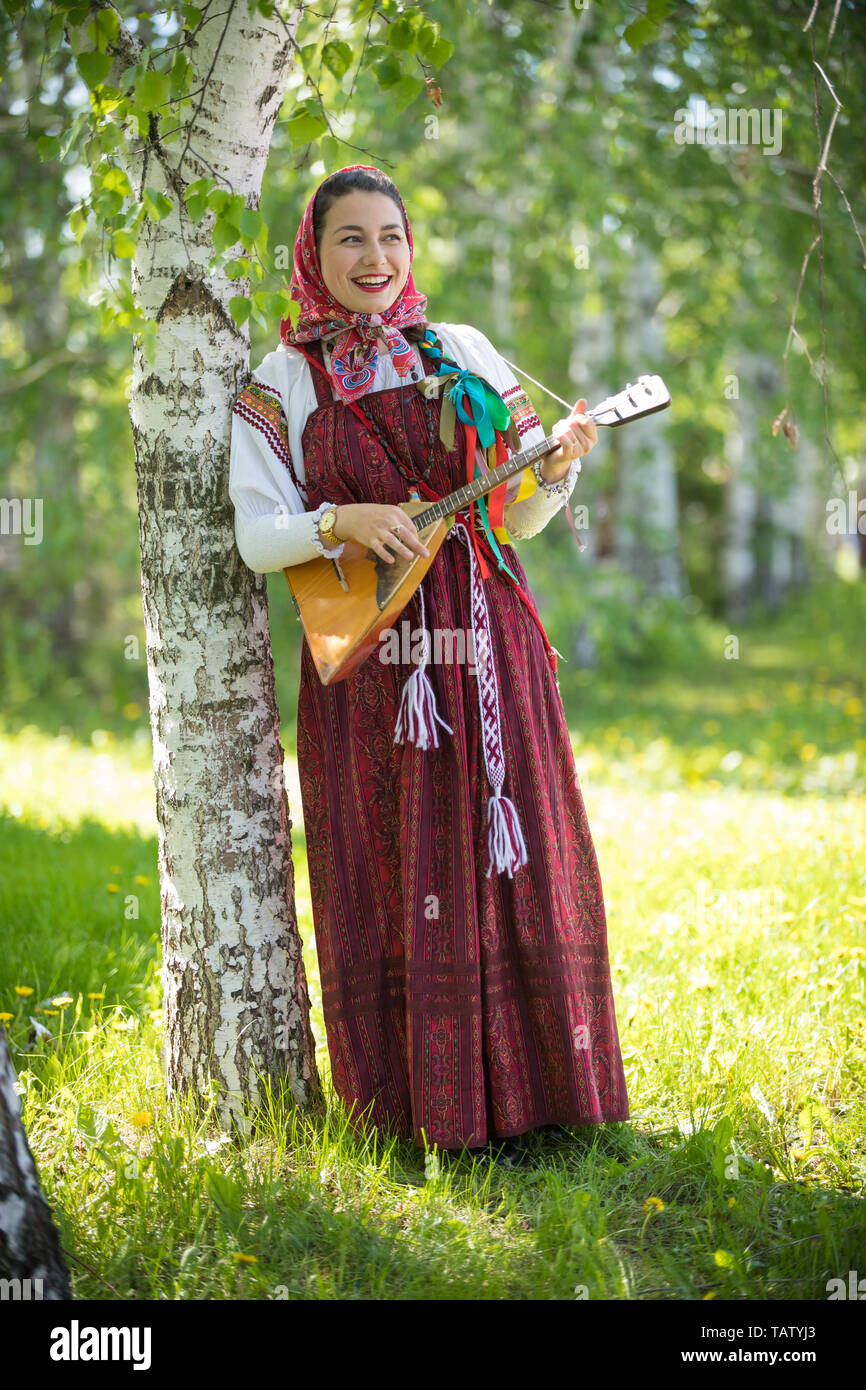 Lächelnde junge Frau in der traditionellen russischen Kleidung stehen im Wald und spielt Balalaika, vertikaler Stockfoto