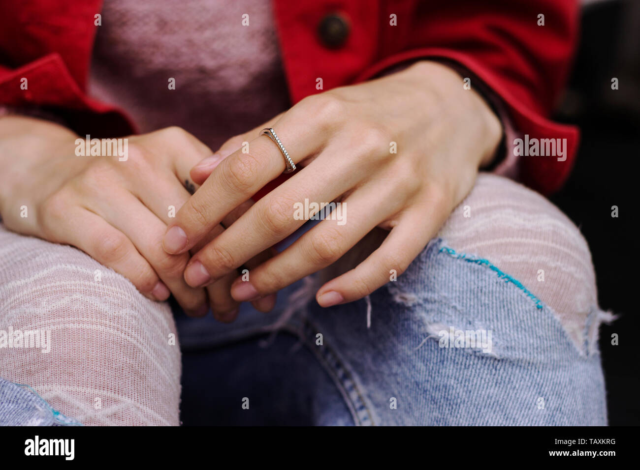 Frau sitzt gekreuzten Beinen mit den Händen in der Runde warten. Zerrissene Jeans und rote Jacke. Berühren Hände besorgt/Sad Stockfoto