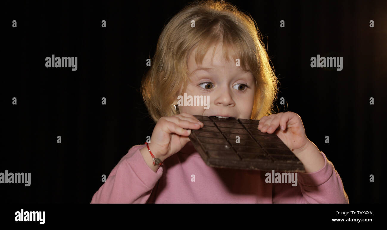 Attraktive Kind Essen einer riesigen Block von Schokolade. Süße blonde Mädchen beißen eine Schokolade. Braune Augen. Hübsches kleines Kind, 3-4 jährige blonde Mädchen. Schwarzer Hintergrund Stockfoto