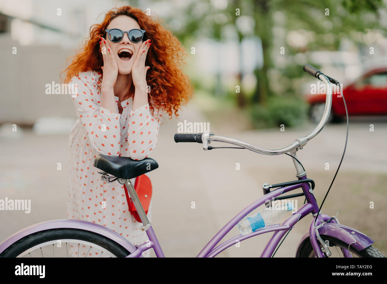 Fröhliche Frau urban Biker trägt Sonnenbrille und Kleid, mit luxuriösen scharfe foxy Haar, hält die Ellbogen auf dem Sattel des Fahrrades, verbringt Freizeit im OP Stockfoto