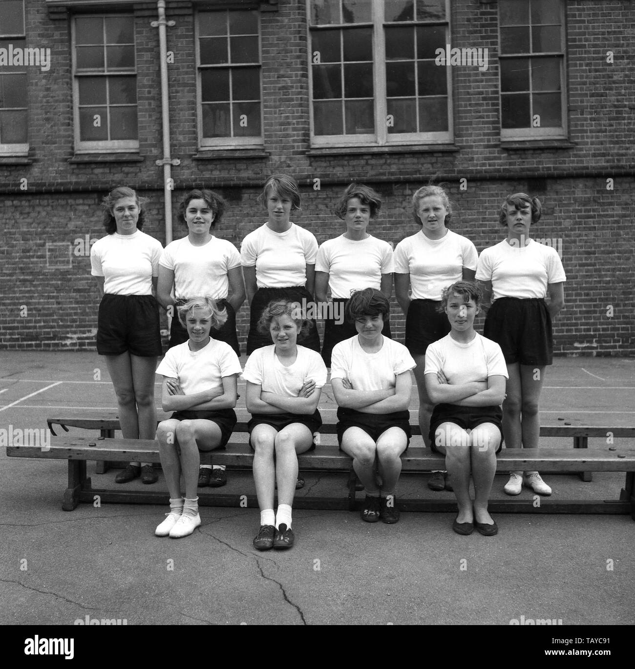 1950, historische, Teenager Sekundärschoolgirls für ein Bild in der PE-kit Posing - einfache weiße T-Shirt, schwarze Shorts und tragen plimsols, außerhalb ihrer innerstädtischen Schulgebäude, England, UK. Stockfoto