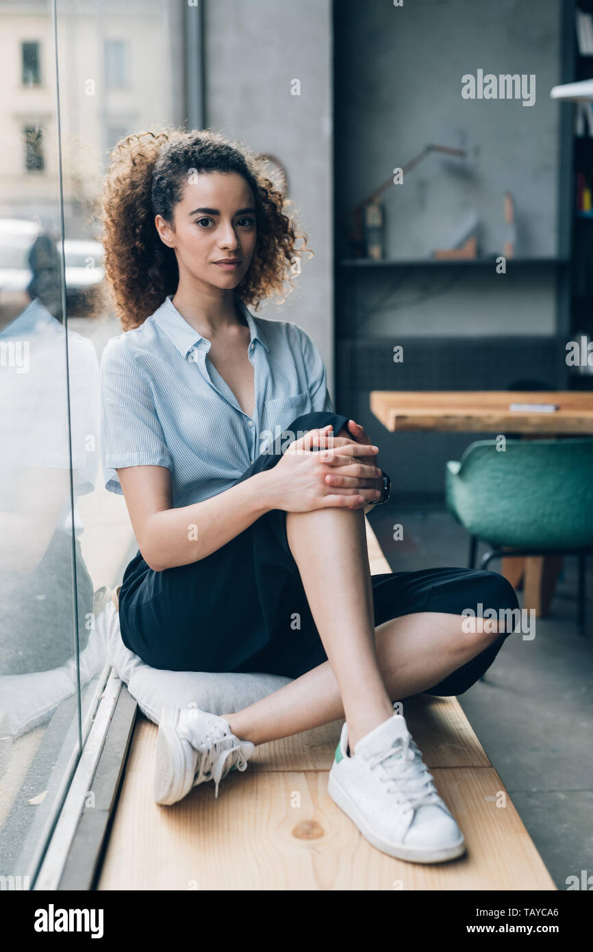 Junge kaukasier Frau mit gekreuzten Beinen in hellen Standort posing und suchen Kamera - Jugendkultur, Emanzipation, neue Generation Stockfoto