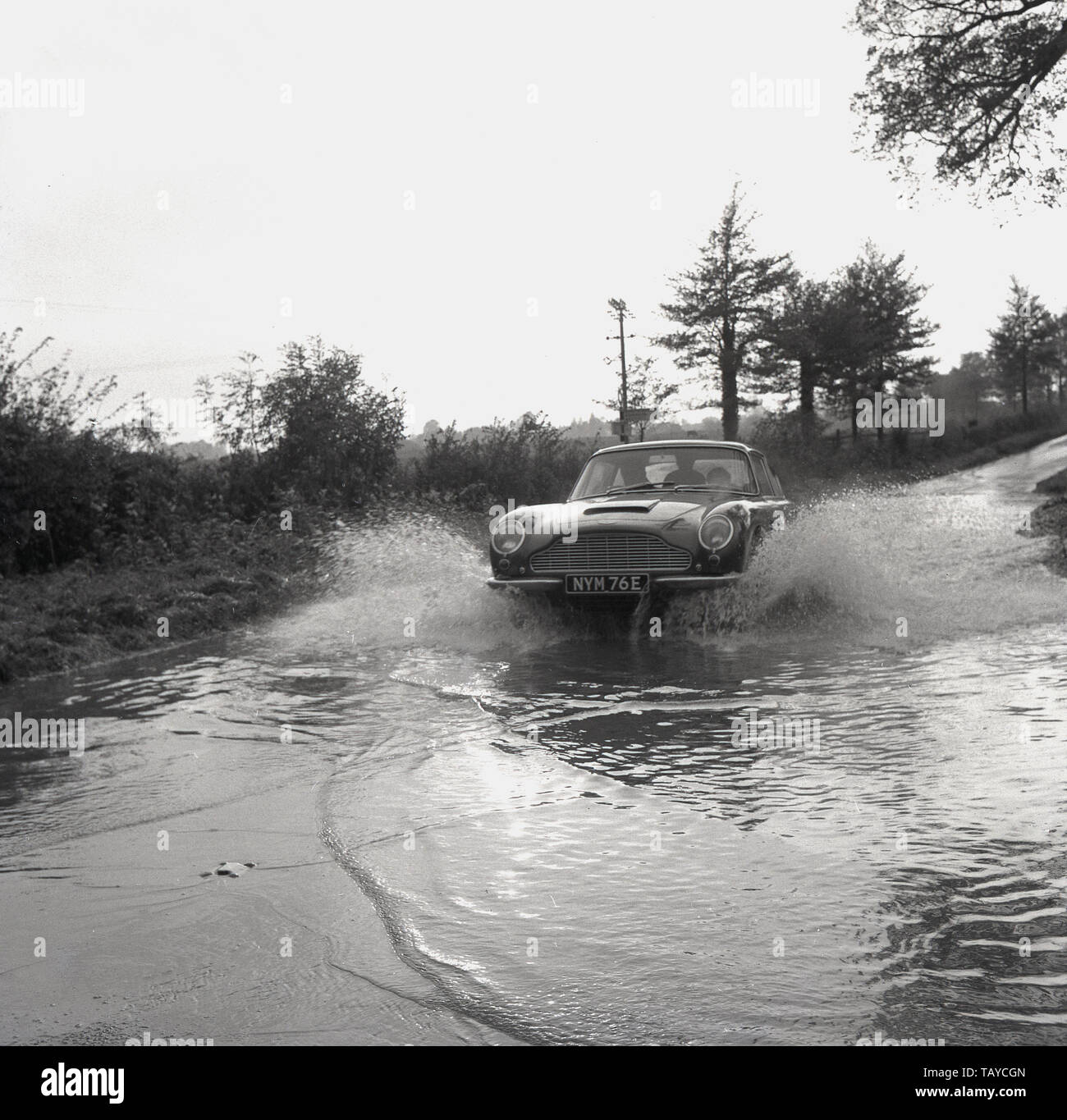 1960, historische, ein Aston Martin Sportwagen erstellt einen splash Wie geht es durch das Wasser auf einem überfluteten Landstraße, Buckinghamshire, England, UK. Stockfoto