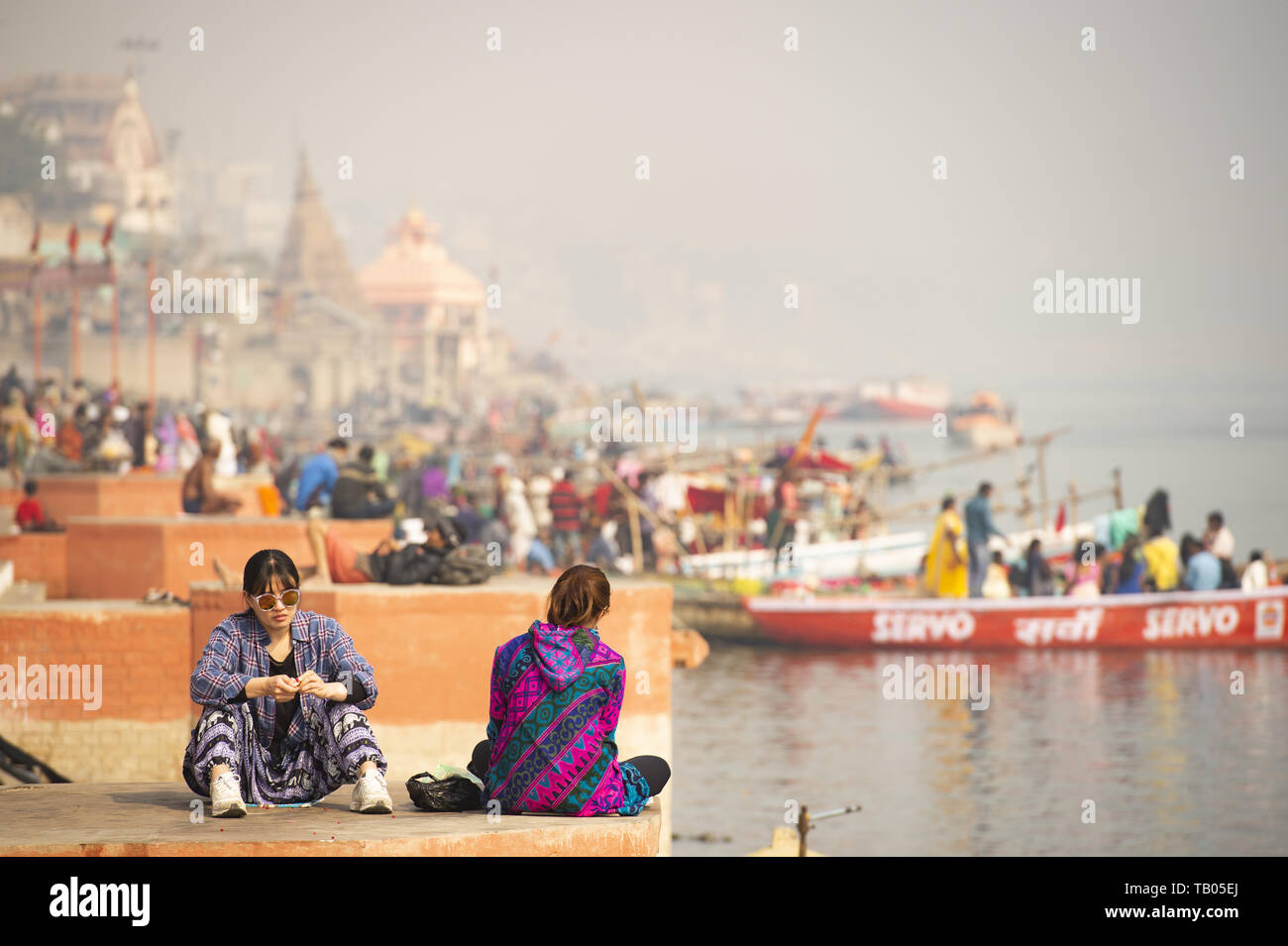 Zwei chinesische Touristen sitzen auf einer Ghat in Varanasi. Manikarnika Ghat (brennende Ghat) im Hintergrund. Varanasi, Indien. Stockfoto