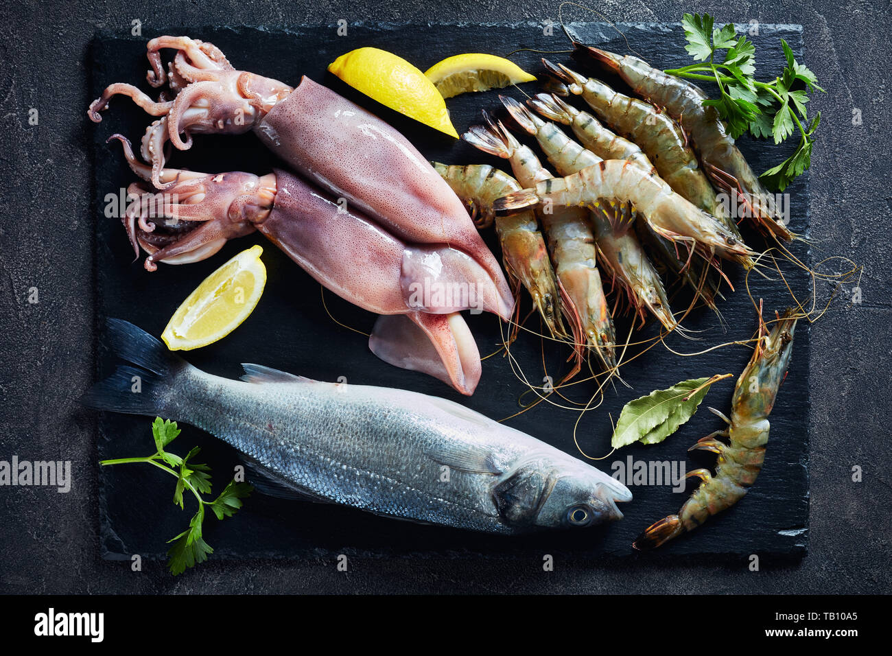 Wolfsbarsch, Garnelen, Tintenfisch, Zitronenscheiben, Gewürze und Kräuter auf einer Schiefertafel Platte auf einer konkreten Tabelle, Ansicht von oben, Nahaufnahme, flatlay Stockfoto