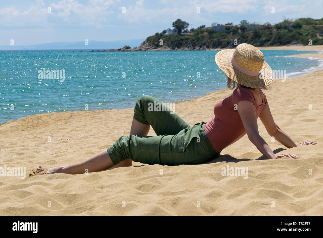 Frau mit casual Outfit und Strohhut sitzend auf Sandstrand und das Meer bewundern. Sommer Urlaub Konzept Stockfoto