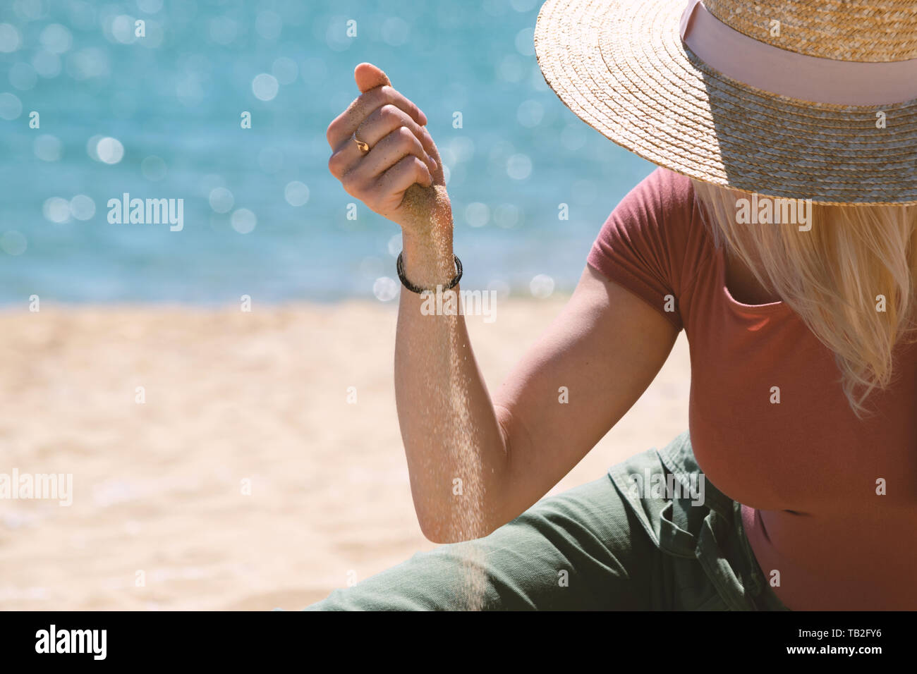 Frau mit casual Outfit und Strohhut sitzend auf Sandstrand vor blauem Meer und Sand durch ihre Hände fließt. Sommer Urlaub Konzept Stockfoto
