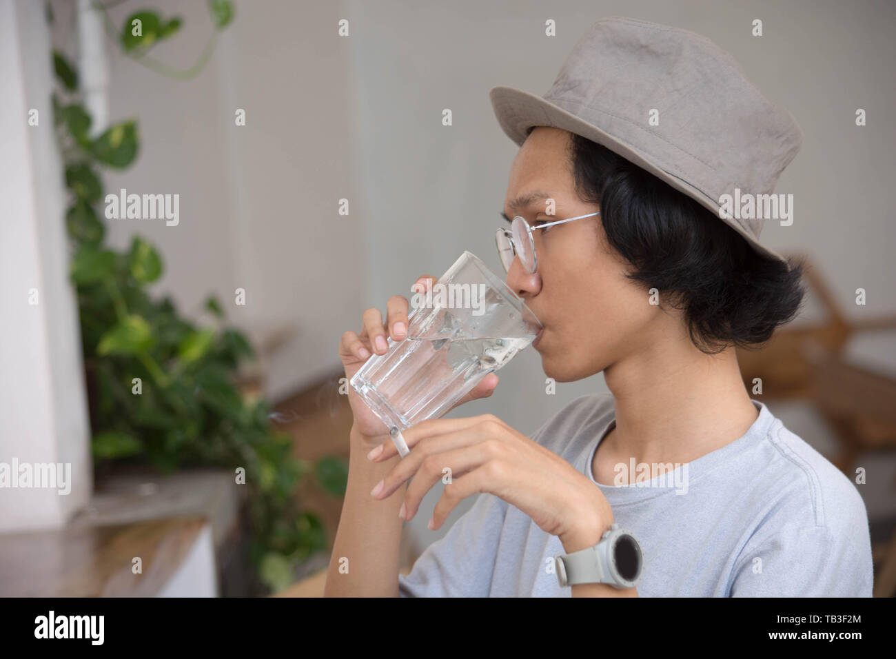 Ein asiatischer Mann mit Fedora Hut und Brille rauchen und trinken Glas Wasser im Cafe Stockfoto