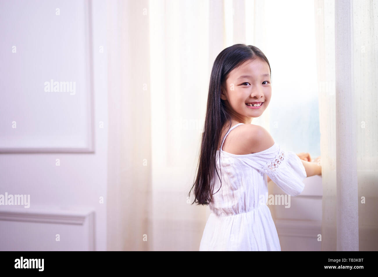 Schöne kleine asiatische Mädchen mit langen schwarzen Haaren stand vor der windwo Drehen in die Kamera lächeln Stockfoto