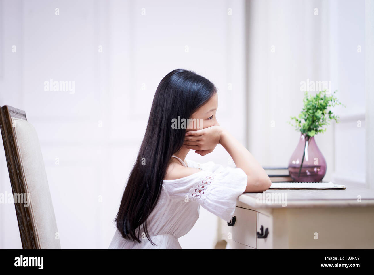 Traurigen kleinen asiatischen Mädchen mit langen schwarzen Haaren sitzen am Schreibtisch in Ihrem Zimmer Denken mit der Hand am Kinn Stockfoto