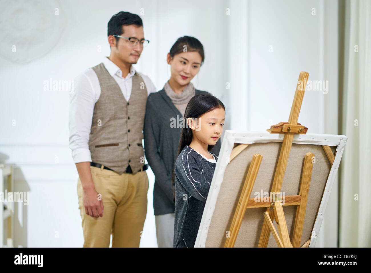 Schöne kleine asiatische Mädchen mit langen schwarzen Haaren und einem Gemälde auf Leinwand, während die Eltern hinter dem Ständigen beobachten. Stockfoto