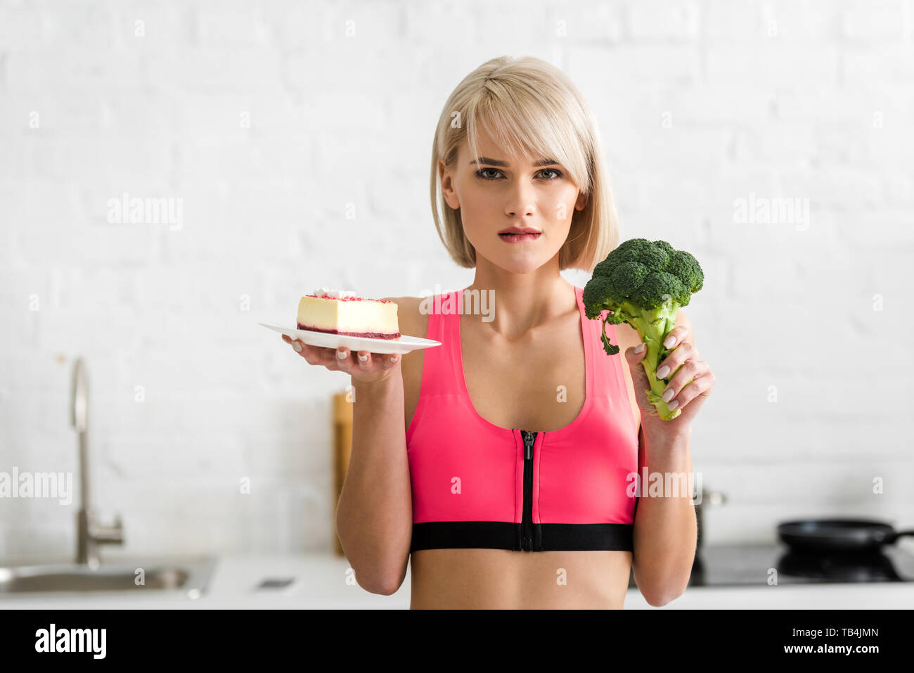 Blonde Mädchen halten Grüne Brokkoli und Untertasse mit süßer Kuchen beim Beißen Lippe Stockfoto