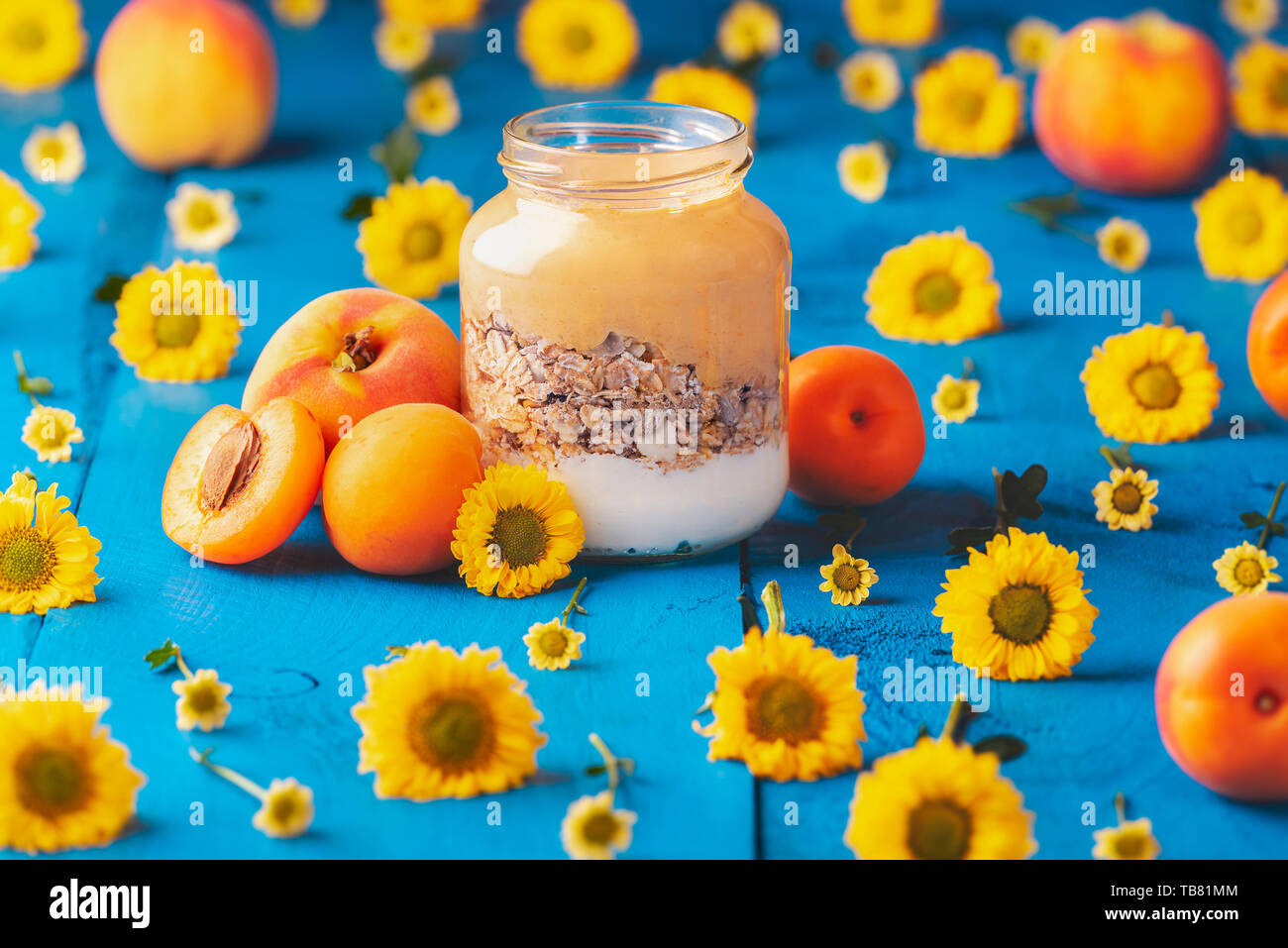 Glas mit Pfirsich Joghurt und Müsli, von Pfirsichen und gelbe Blumen umgeben, auf einem blauen Holztisch. Diät Frühstück. Bereit, gesunde Lebensmittel zu essen. Stockfoto