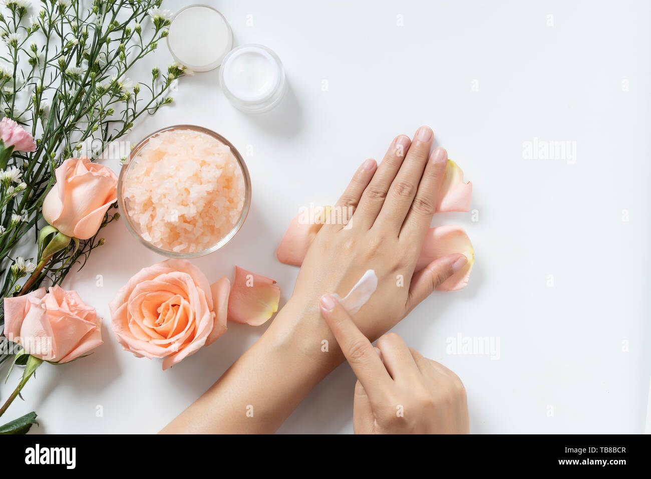 Natürliche Hautpflege Konzept. Frau Weiß Creme auf die Hände auftragen auf weißem Hintergrund mit Jar von kosmetischen Creme, Salz Spa Peeling, rosa und weißen Blüten Stockfoto