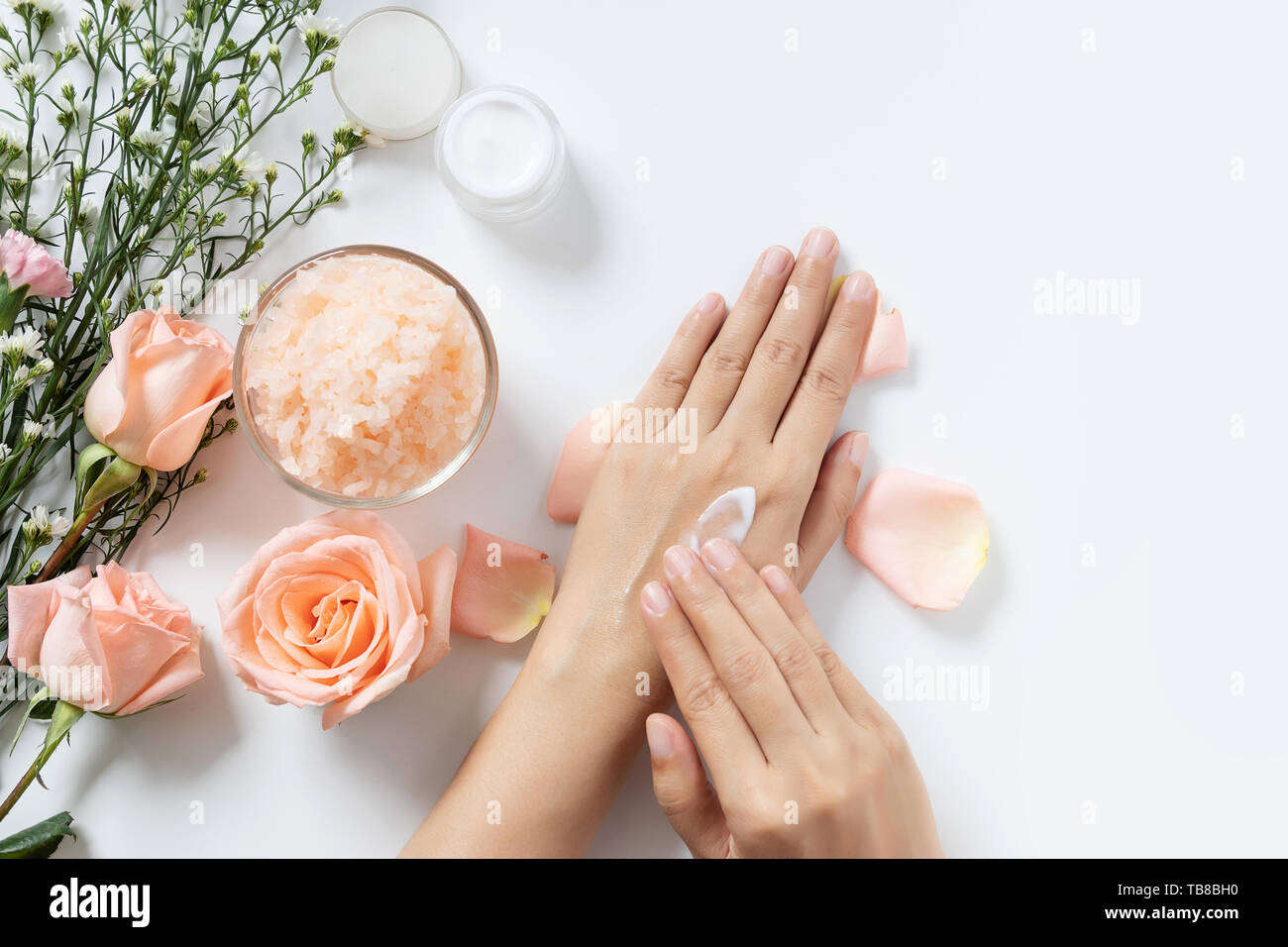 Natürliche Hautpflege Konzept. Frau Weiß Creme auf die Hände auftragen auf weißem Hintergrund mit Jar von kosmetischen Creme, Salz Spa Peeling, rosa und weißen Blüten Stockfoto