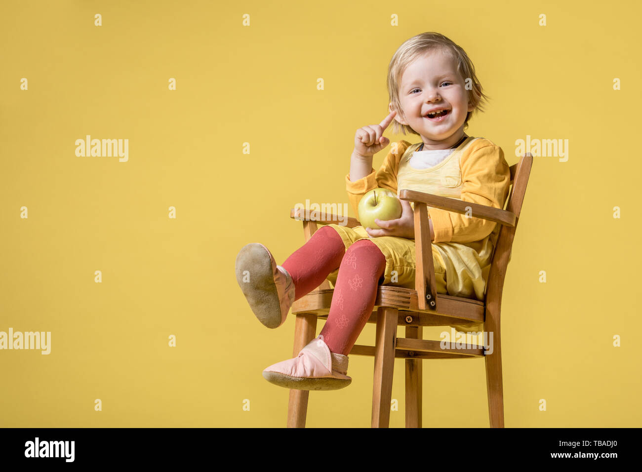 Junge Blonde Mädchen im gelben Kleid, Sitzen auf den Kinderstuhl und fröhlich lachen, lächeln und Essen Green Apple auf gelbem Hintergrund Stockfoto