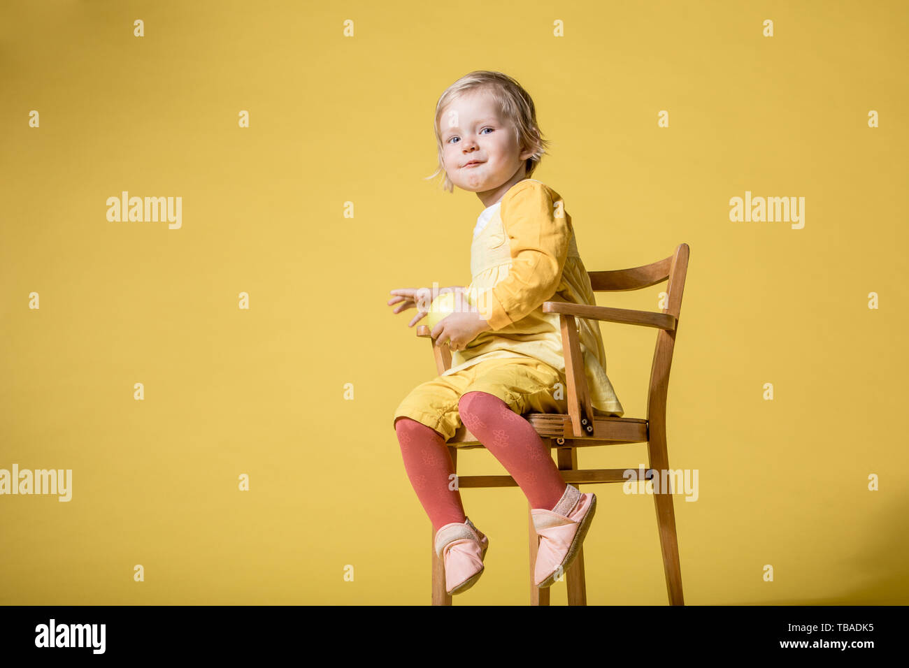 Junge Blonde Mädchen im gelben Kleid, Sitzen auf den Kinderstuhl und fröhlich lachen, lächeln und Essen Green Apple auf gelbem Hintergrund Stockfoto
