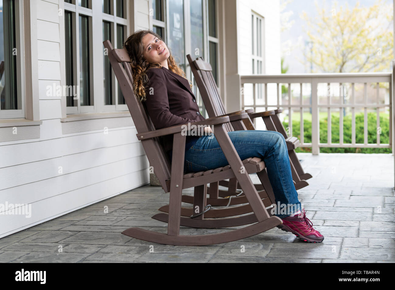 Veranda vor dem Haus mit Jungen glücklich Frau sitzt auf der braunen Schaukelstuhl in der traditionellen amerikanischen Haus Stockfoto
