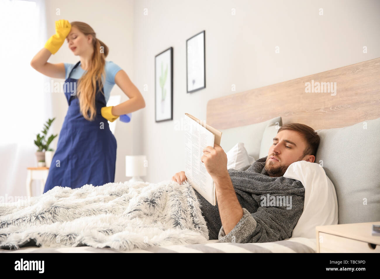 Faul mann lesen Zeitung, während seine müde Frau tun Chores zu Hause Stockfoto