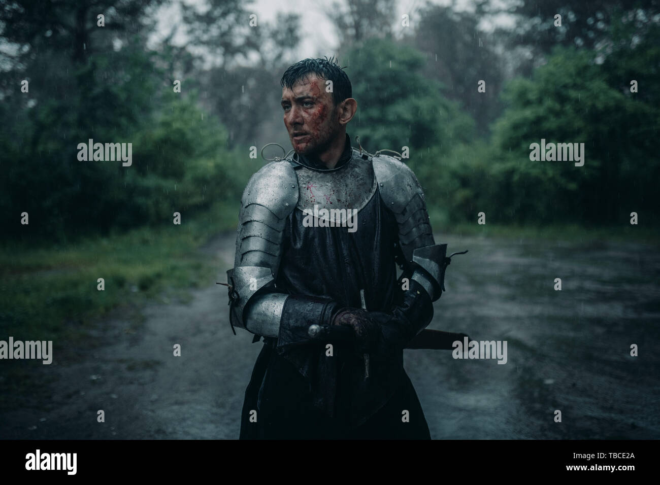 Ein junger Mann steht in der Bild einer mittelalterlichen Ritter in Rüstung und mit dem Schwert in der Hand im Regen. Stockfoto