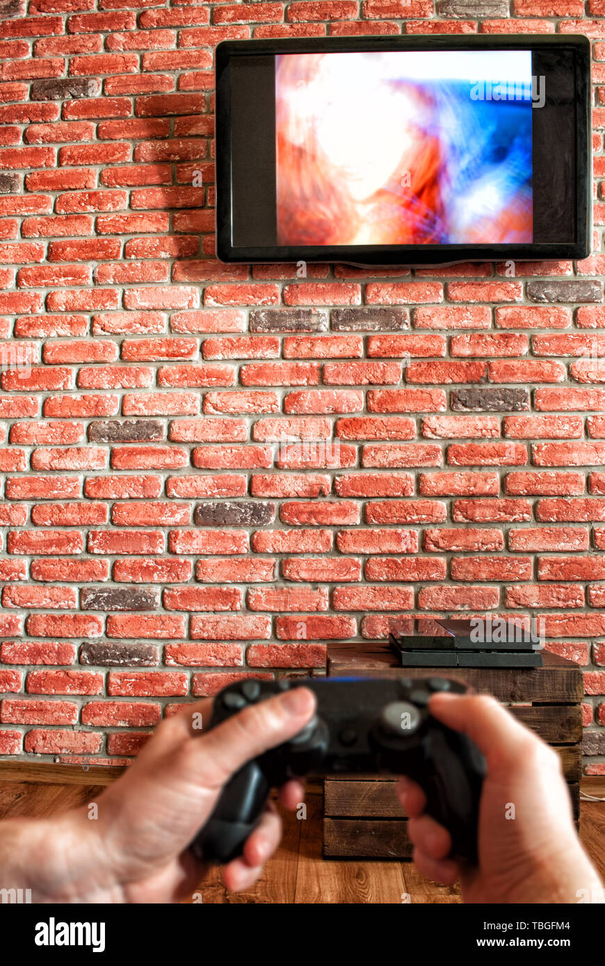 Brick Wall mit TV und Fernbedienung von der Spielekonsole in den Händen im Vordergrund. Konzentrieren Sie sich auf die Wand mit TV Stockfoto