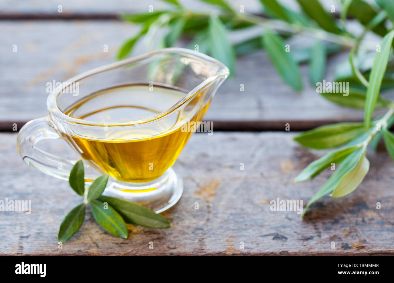 Olivenöl in Glaskanne mit Olivenbaum Zweige auf Holz- Hintergrund. Nahaufnahme Stockfoto
