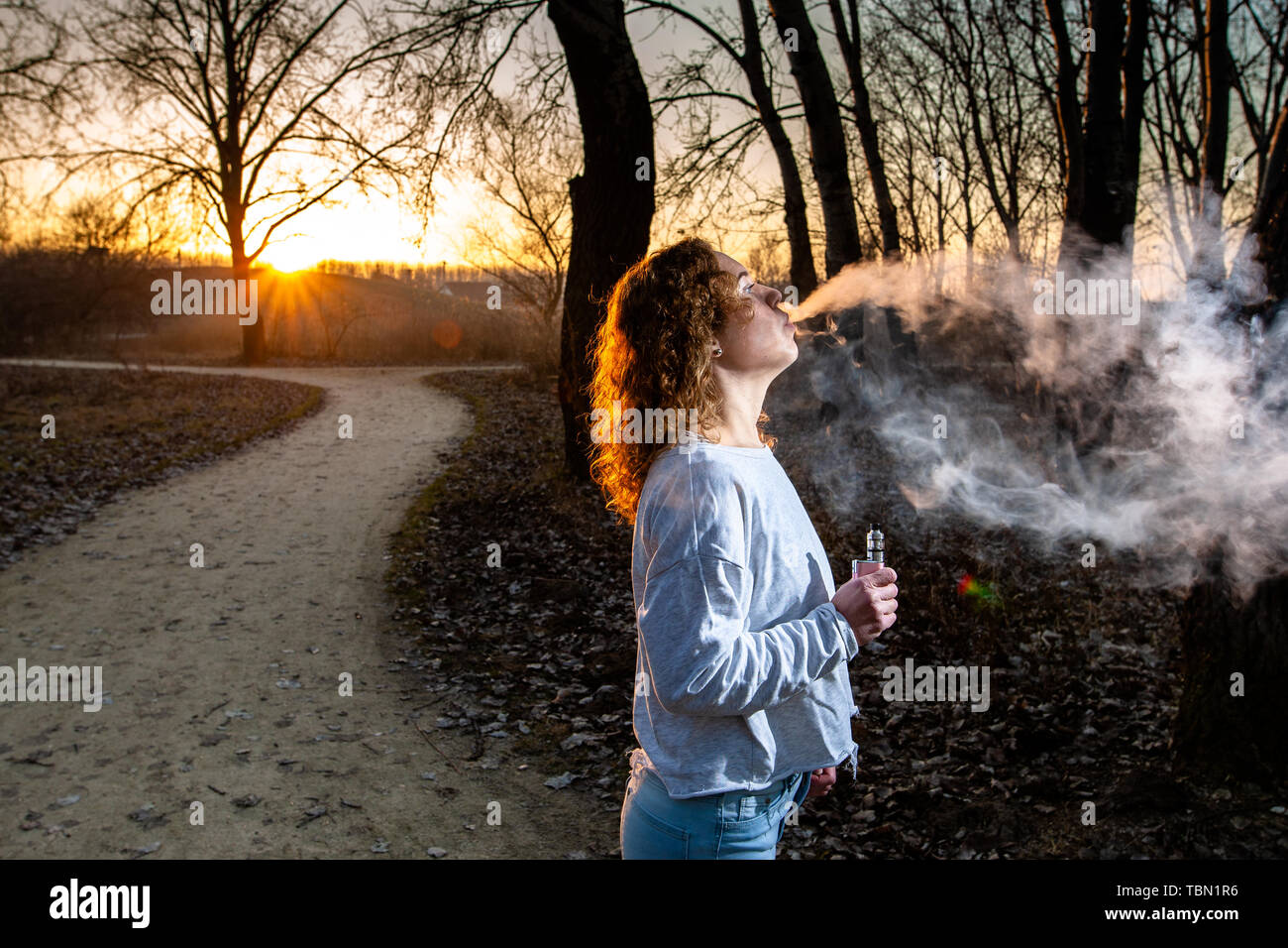 Junge Frau rauchen elektronische e-Zigarette im Park an einem sonnigen Tag bei Sonnenuntergang mit Rauch. Stockfoto