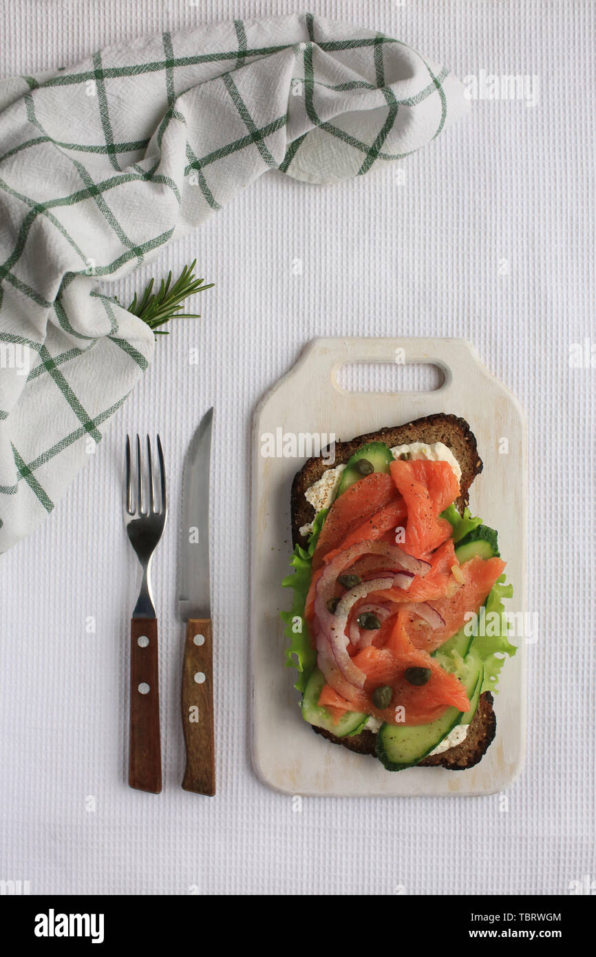 Big leckere Smorrebrod mit Lachs, Grün vegatbles, Zwiebeln, Kapern, Gurken, Käse, Pfeffer und Roggenbrot Festlegung auf weißem Handtuch auf Holzbrett. Messer und Gabel mit Holzgriffen sind in der Nähe. Stockfoto