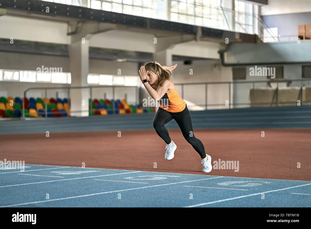 Unglaublich Mädchen in einer schwarz-orange T-Shirt grau mit Dunklen Hosen und Turnschuhe ist auf Startposition auf der Laufstrecke Nummer 5 an der Indoor Stadium. Stockfoto