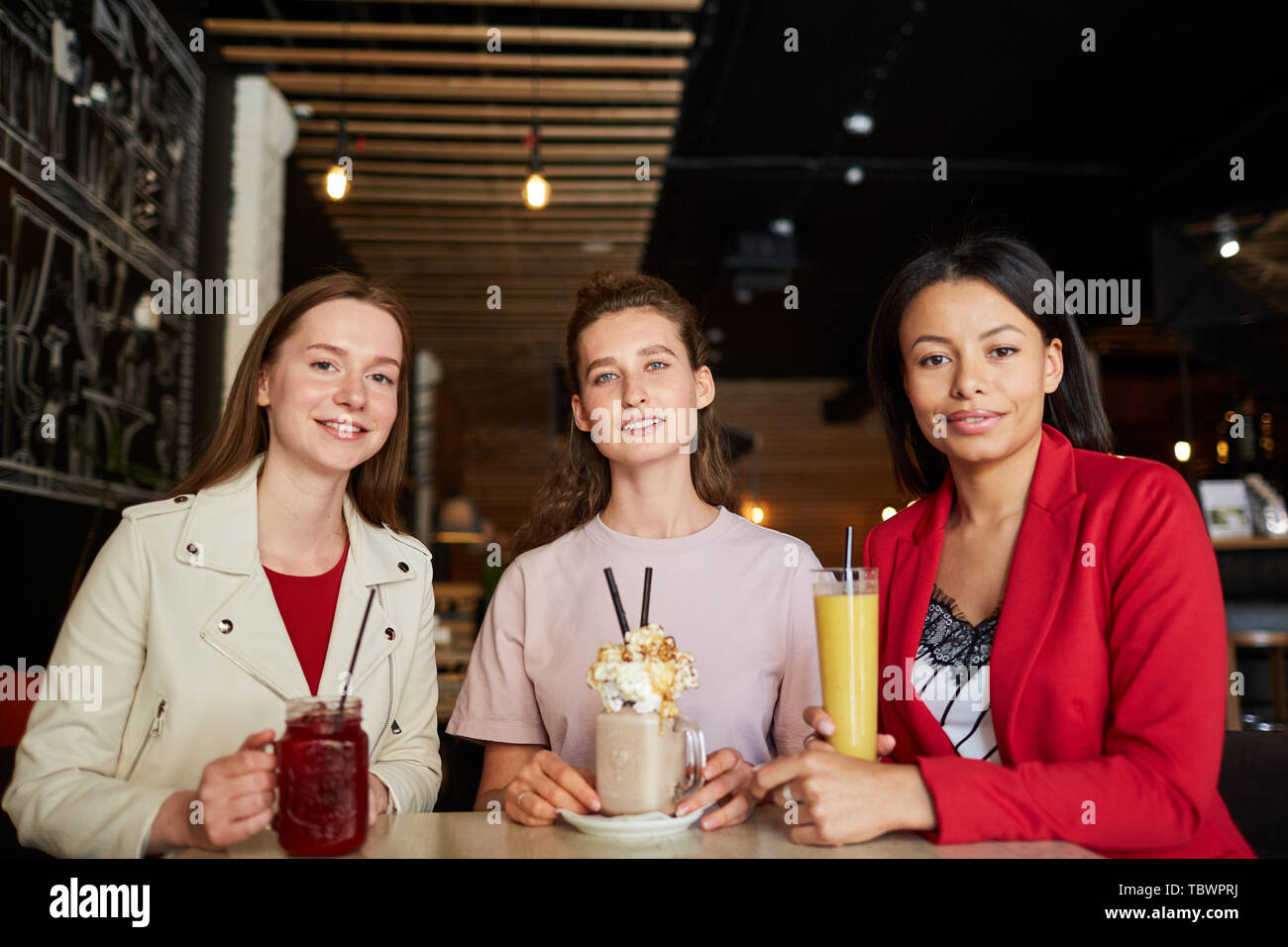 Positive hübsche junge multi-ethnischen Frauen in casual Outfits am Tisch saßen und an Kamera und genießen Sie süße Getränke im Café Stockfoto