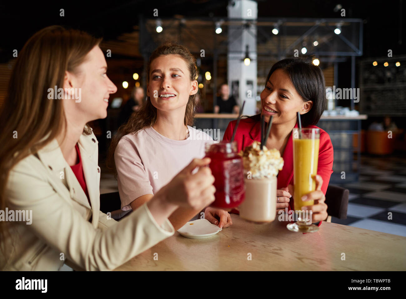 Glückliche unbeschwerte junge multi-ethnischen Frauen sitzen am Tisch und sagt Witz Toast mit süße Getränke, während Spaß im Cafe Stockfoto