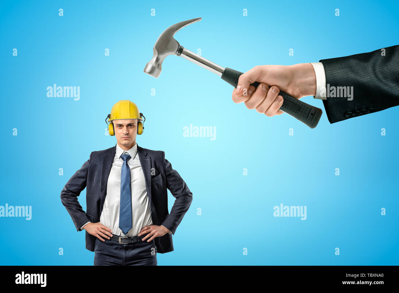 Unternehmer im gelben Schutzhelm mit Gehörschutz, auf den Hüften, und die große Hand, die Hammer auf den Kopf. Stockfoto
