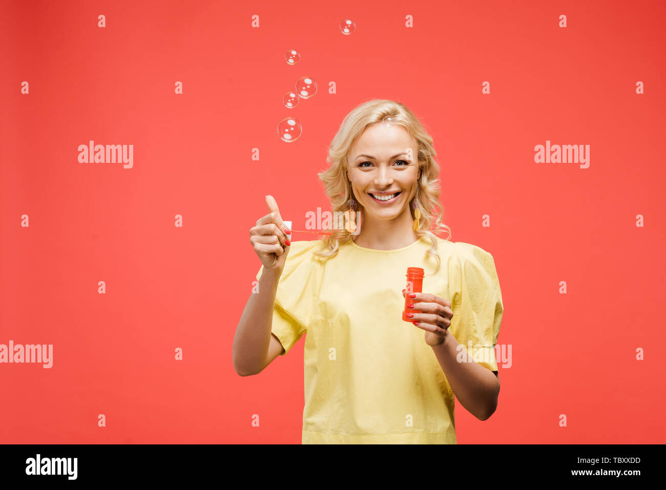 Fröhliche blonde Frau mit Flasche in der Nähe von Seifenblasen und Daumen hoch, rot Stockfoto
