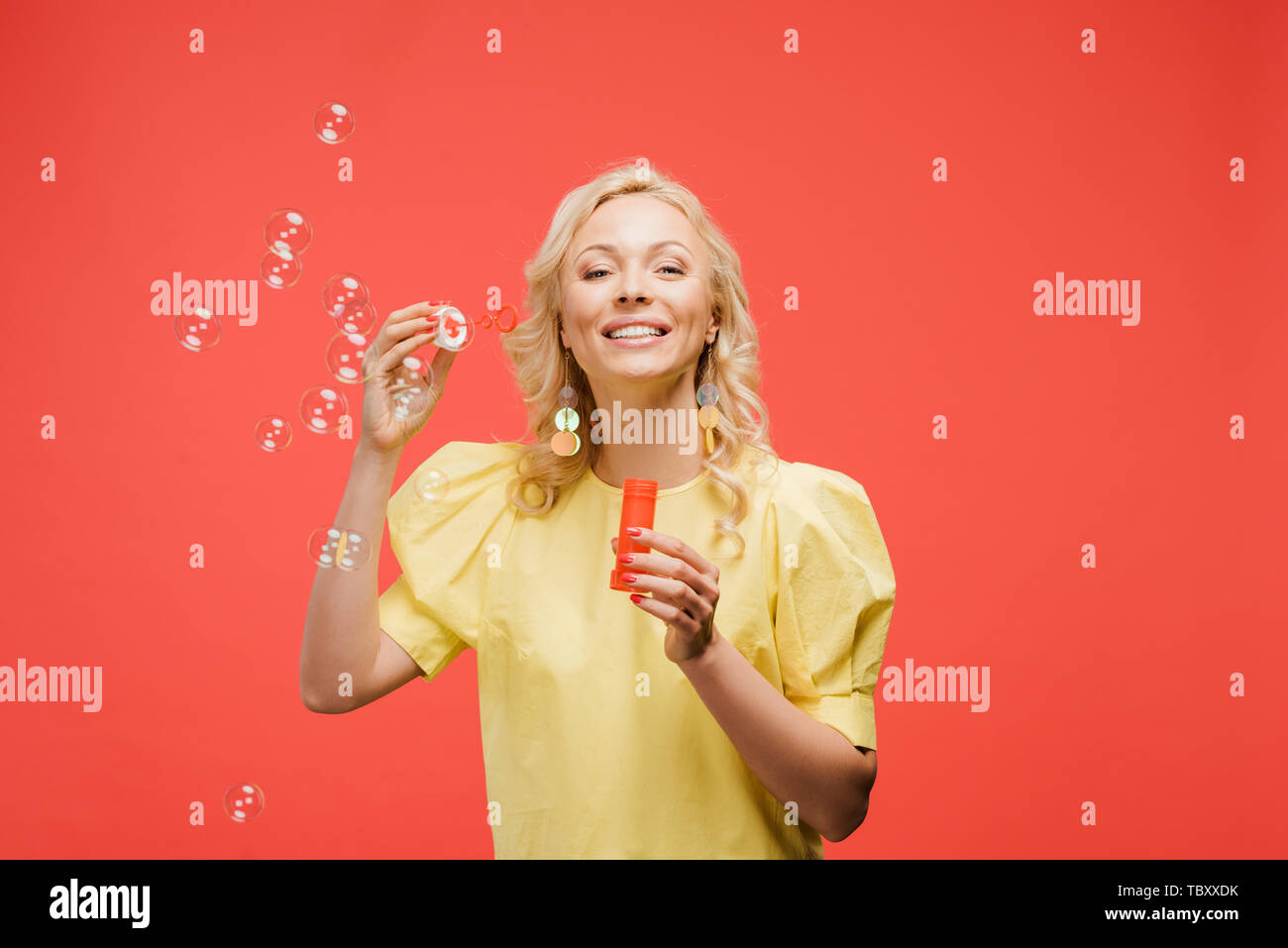 Fröhliche blonde Frau mit Flasche in der Nähe von Seifenblasen und lächelnd auf Rot Stockfoto
