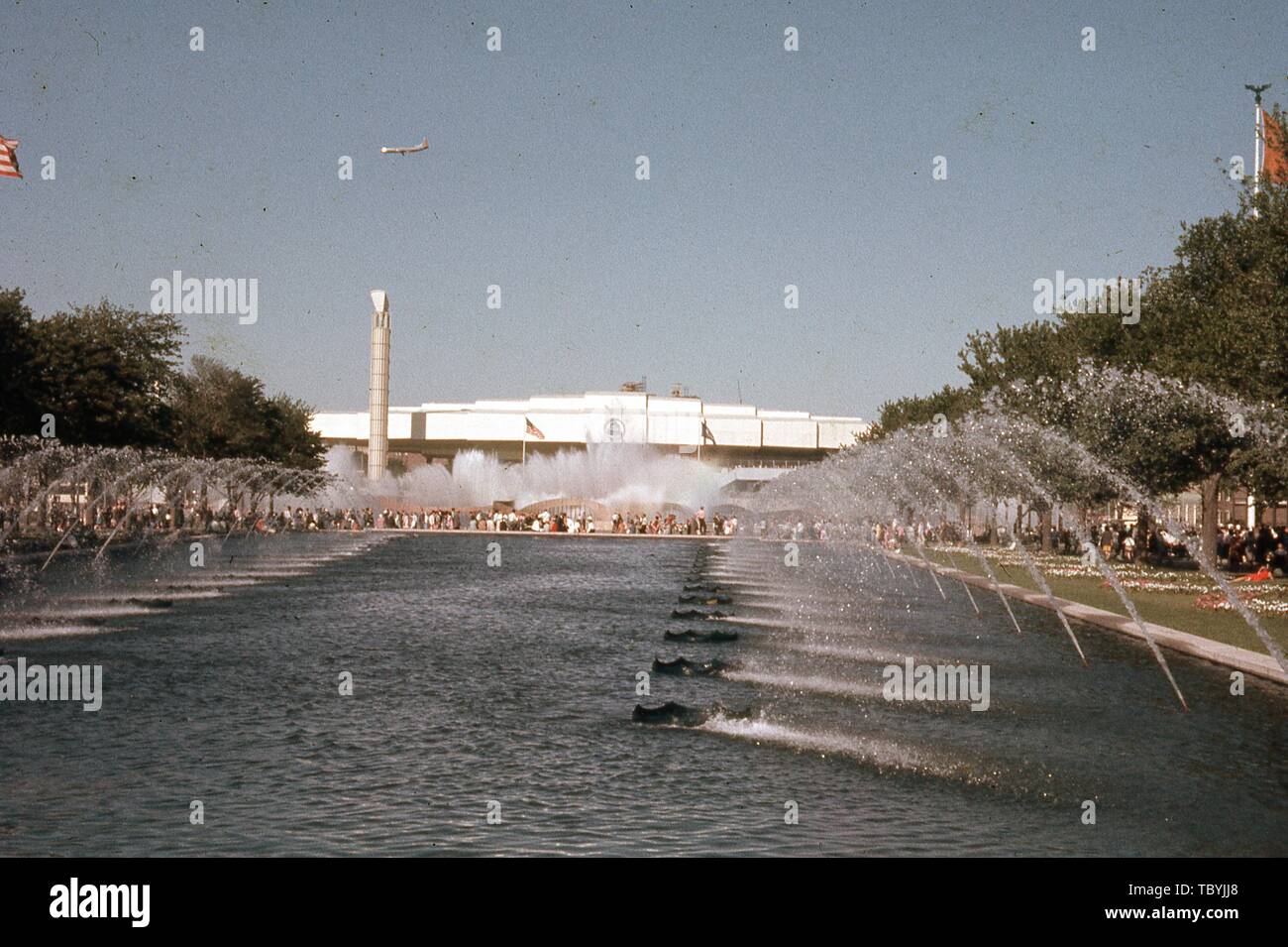 Long Shot, an einem sonnigen Tag, der Massen in der Nähe von einem Brunnen und einem Pavillon, mit einem Flugzeug, Overhead, 1964 in New York World's Fair, Flushing Meadows Park, Queens, New York, Juni, 1964. () Stockfoto
