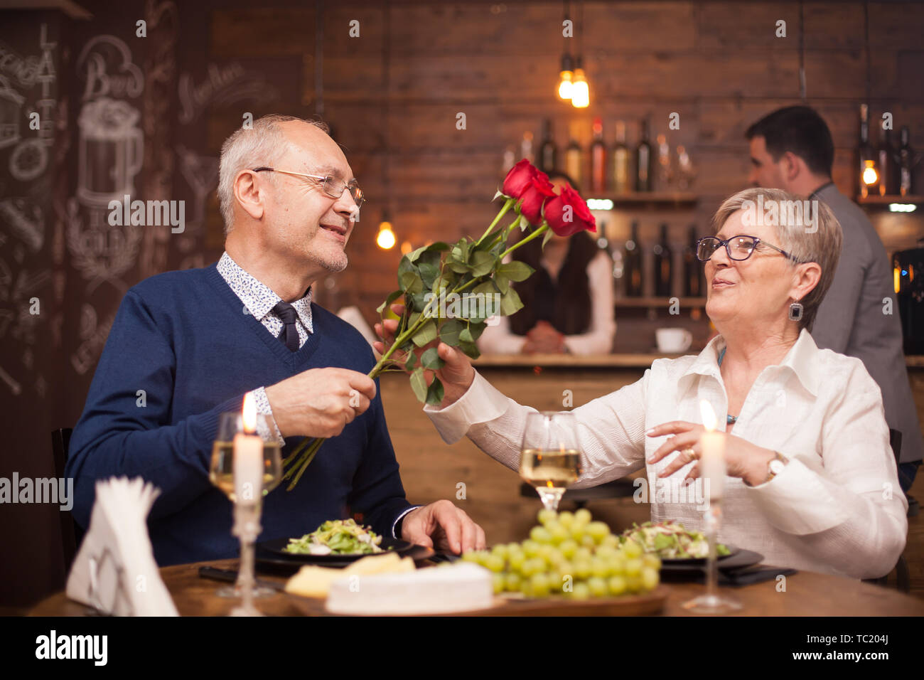 Frau in den 60er Jahren gerne Rosen von ihrem Ehemann beim Abendessen zu erhalten. Senior paar Datum. Frischen Weintrauben. Freundliche alte Paar. Stockfoto