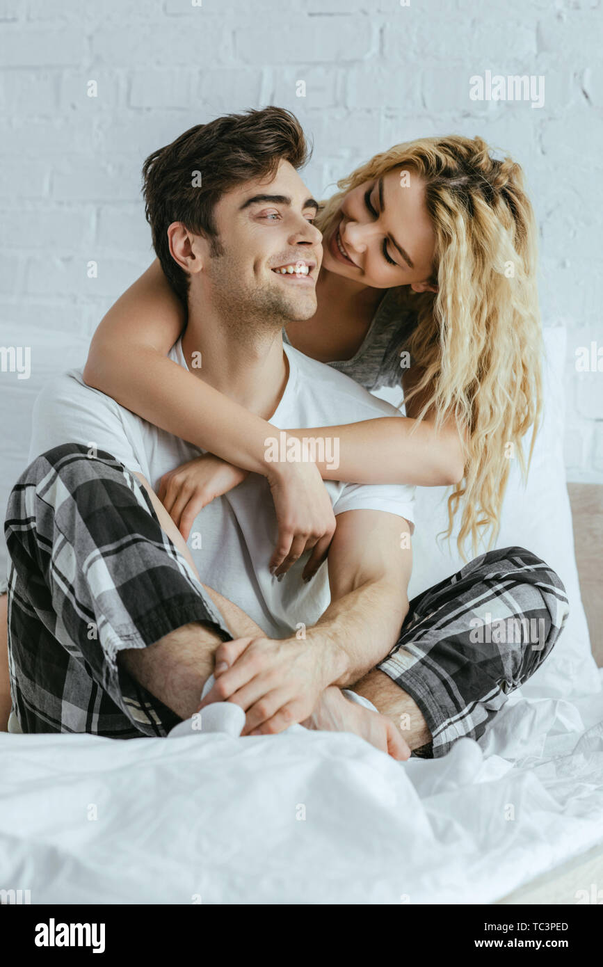 Attraktive blonde Frau umarmt glücklich Freund saß auf dem Bett Stockfoto