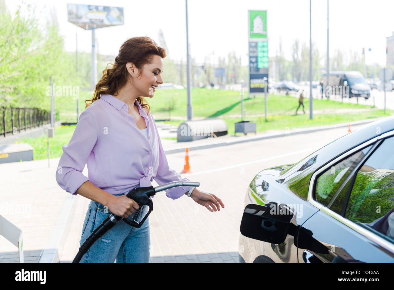 Fröhliche Frau, die Kraftstoffpumpe in der Nähe von Schwarzen Auto an der Tankstelle Stockfoto