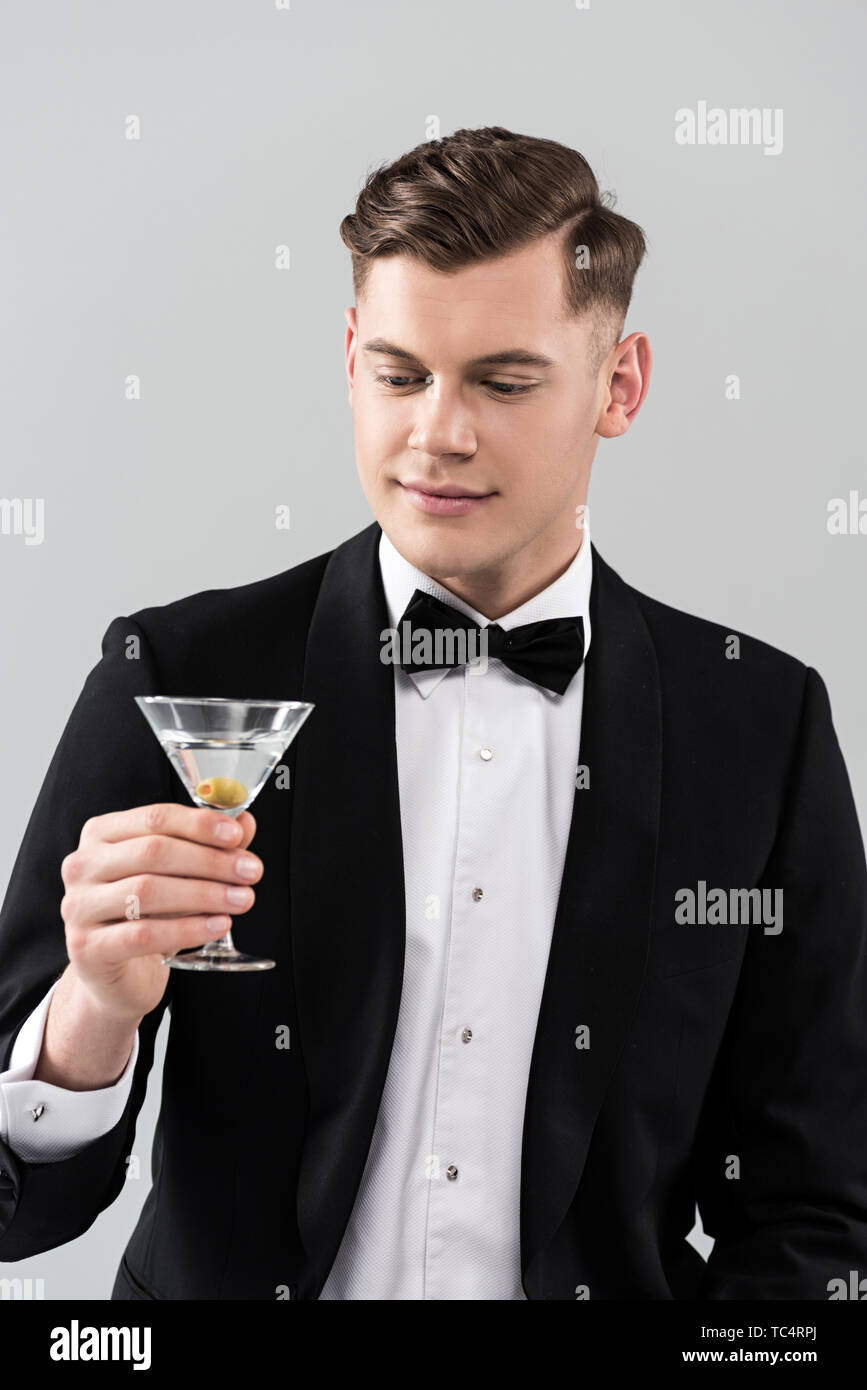 Lächelnden jungen Mann in formale Abnutzung mit Bow Tie holding Glas cocktail isoliert auf Grau Stockfoto
