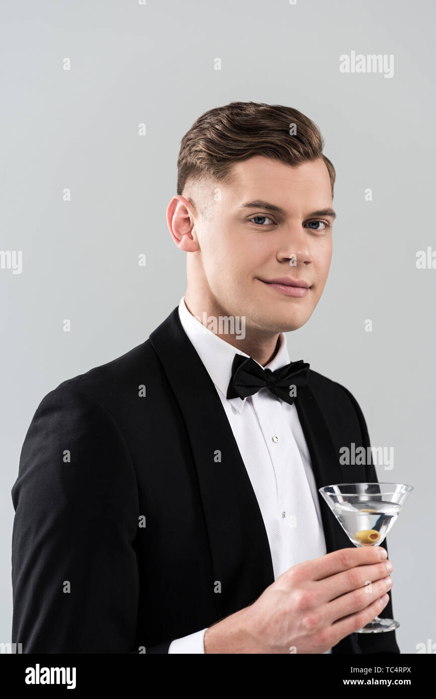 Lächelnden jungen Mann in formale Abnutzung mit Bow Tie holding Glas cocktail isoliert auf Grau Stockfoto