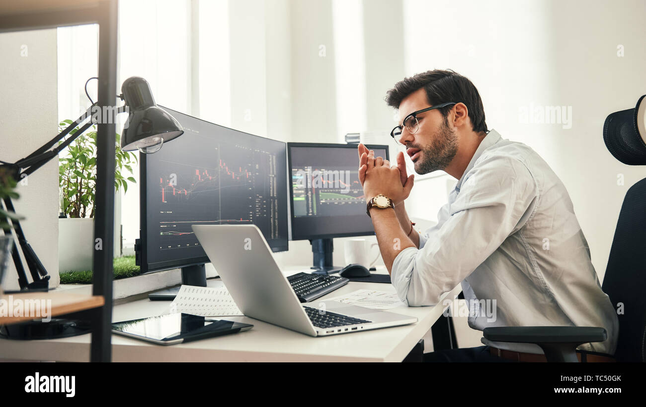 Trading Strategie. Erfolgreiche junge Unternehmer in Brillen zu analysieren trading Charts auf Computerbildschirmen in Während in seinem modernen Büro zu sitzen. Börsenmakler. Forex Markt. Handel Konzept Stockfoto